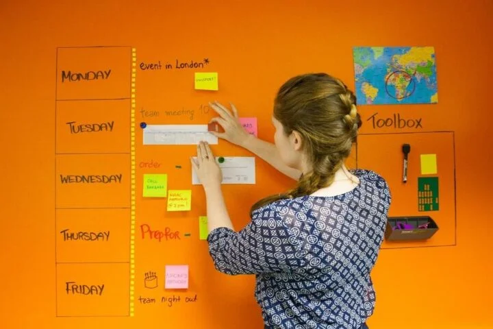 A girl is placing a paper with magnets on a magnetic whiteboard paint wall with a world map alongside a weekly summary of the London event on the wall