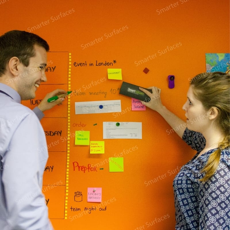 Two professional colleagues collaborating at vibrant orange magnetic whiteboard paint clear wall
