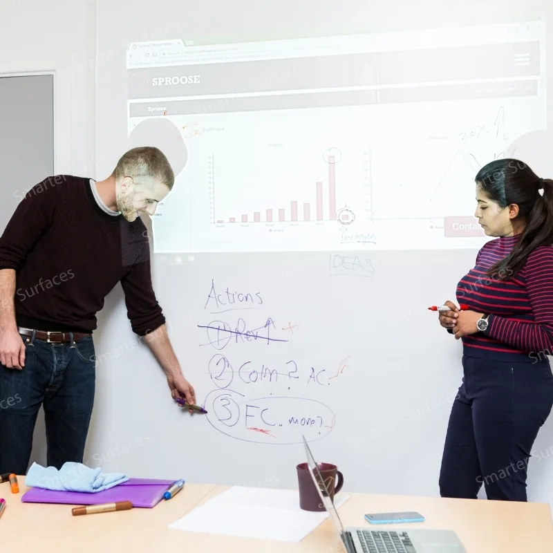 Two colleagues collaborating at matte whiteboard paint wall. Man writing action items and deadlines while woman observes with marker.
