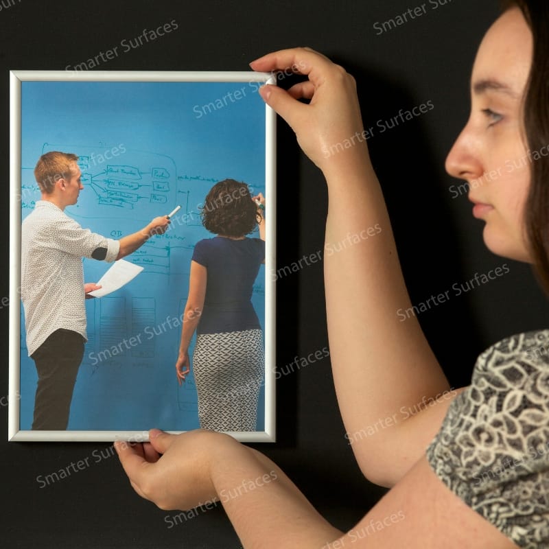 Woman hanging framed photograph of office meeting on black strong magnetic paint wall without using nails or hooks
