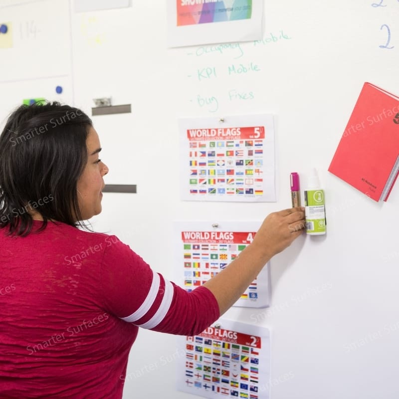 Woman in red shirt writing on transparent magnetic paint whiteboard wall in educational setting.