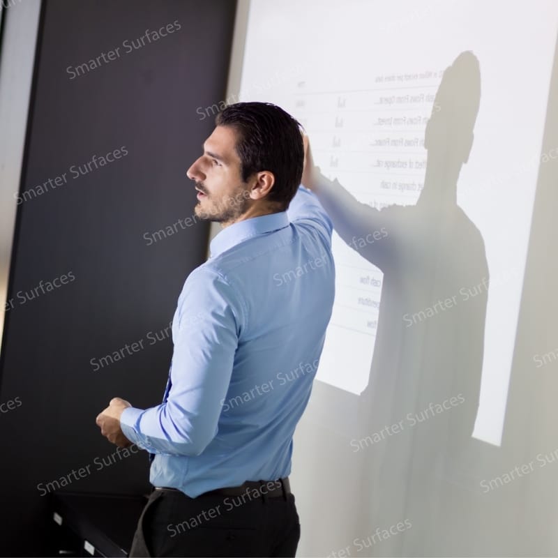 A man giving a presentation while pointing at a projected display on a smooth white wall coated with projector screen paint.