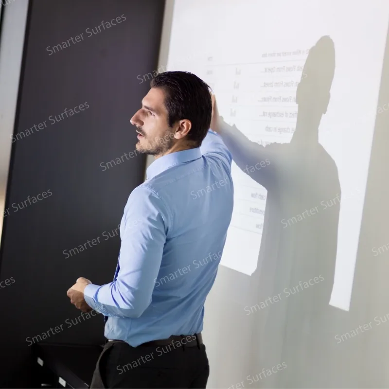 A man giving a presentation while pointing at a projected display on a smooth white wall coated with projector screen paint.