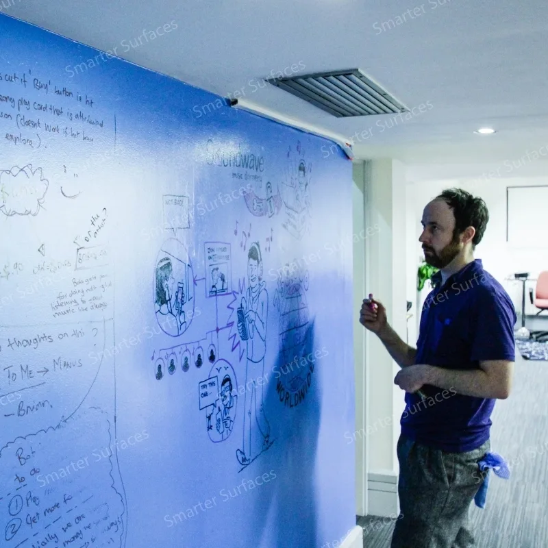 A man is writing on a large blue wall painted with dry erase whiteboard paint.