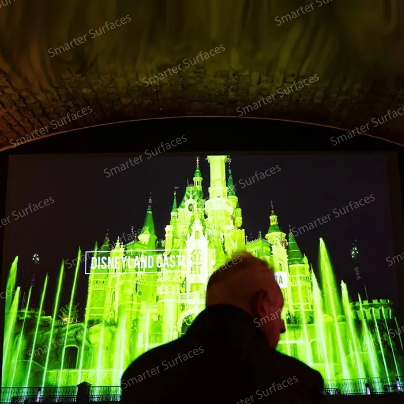 Person viewing castle projection illuminated in bright green with light beams outdoors through silver screen paint projection
